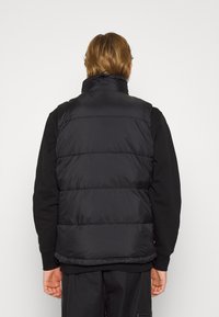 Gilet imbottito nero con colletto alto, sezioni trapuntate visibili e una texture liscia e lucida. Indossato sopra un felpa scura.