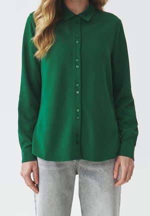 Femme portant une chemise boutonnée à manches longues vert foncé et un jean gris clair, debout devant un fond uni.