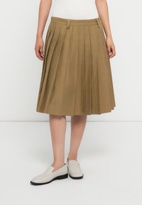 Gonna khaki plissettata realizzata in tessuto testurizzato, con cintura e design che arriva fino al ginocchio. Abbinata a scarpe slip-on di colore chiaro.