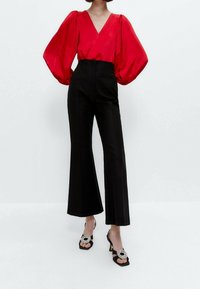 Blusa rossa in satin con maniche a sbuffo, scollatura a V, abbinata a pantaloni neri a vita alta e svasati. I tacchi neri presentano un accento decorativo circolare.