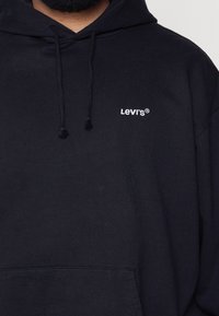 Homme portant un sweat à capuche noir avec le logo Levi's en blanc sur la poitrine gauche et des cordons pendants à la capuche.