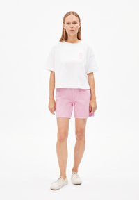 Witte, cropped, kortemouw t-shirt met een roze lint logo; gecombineerd met lichtroze, knielange shorts. Witte sneakers maken de outfit af.