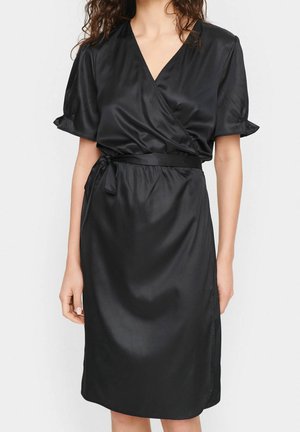 Vrouw draagt een zwarte satijnen wikkeljurk met korte geplooide mouwen en een ceintuur in de taille, staand tegen een eenvoudige lichte achtergrond.
