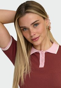 Jeune femme aux longs cheveux blonds portant des boucles d'oreilles en or et un haut côtelé bordeaux avec un col rose, reposant un bras derrière la tête.
