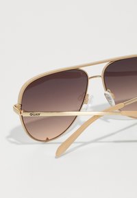 Gafas de sol aviador de metal dorado con lentes marrones degradados, almohadillas nasales transparentes y patas delgadas, con el nombre de la marca "QUAY" en la montura.