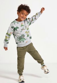 Smiling child balancing on one foot, wearing a gray dinosaur-print sweatshirt, olive green pants, and white sneakers against a white background.