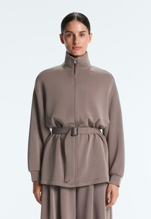 Žena oblečená v taupe bunde s vysokým golierom na zips, s opaskom v páse a v rovnakej sukni, stojaca pred jednoduchým svetlým pozadím.