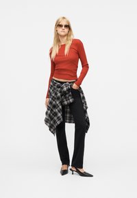 Femme blonde portant des lunettes de soleil, un haut à manches longues couleur rouille, un pantalon noir, des talons noirs, avec une chemise à carreaux noir et blanc nouée autour de la taille.