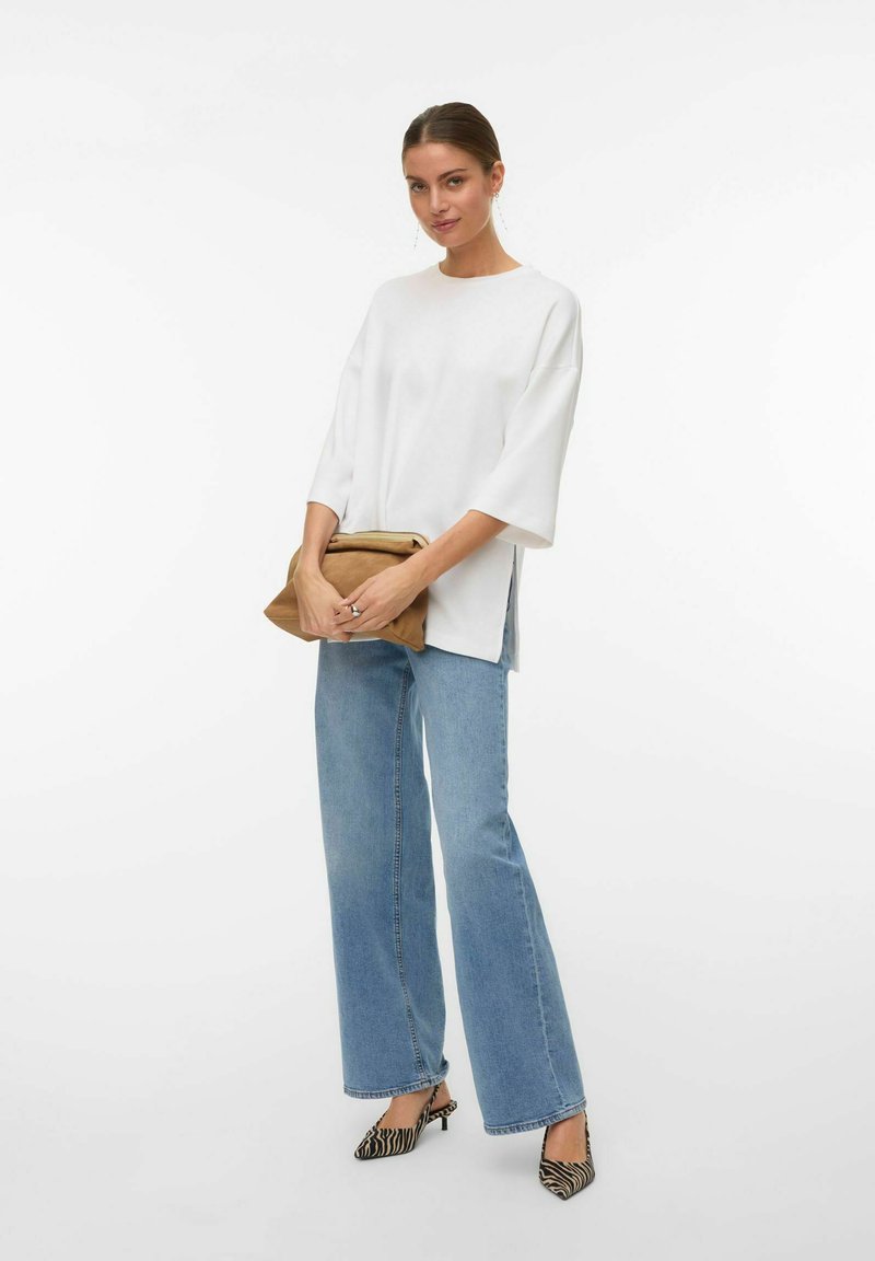Femme portant un haut ample blanc, un jean large bleu clair, des talons imprimés zèbre, tenant une pochette brune pliée, debout sur un fond blanc.