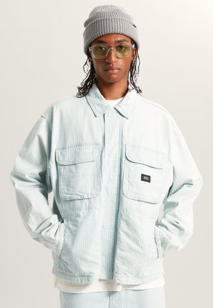 Vaaleansininen raidallinen takki, jossa on kaksi rintataskua ja Vans-logo, valkoisen paidan päällä, yhdistettynä harmaaseen neulepipoon ja kirkkaisiin laseihin.