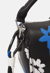 Musta keinonahkainen laukku sinivalkoisilla kukkakuosisilla, jossa on musta karabiini ja teksturoitu kahva. Kompakti, sylinterimäinen muoto.