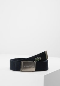 Navy fabric belt featuring a metallic rectangular buckle with engraved "LACOSTE" and a green crocodile logo embroidered on the strap.
