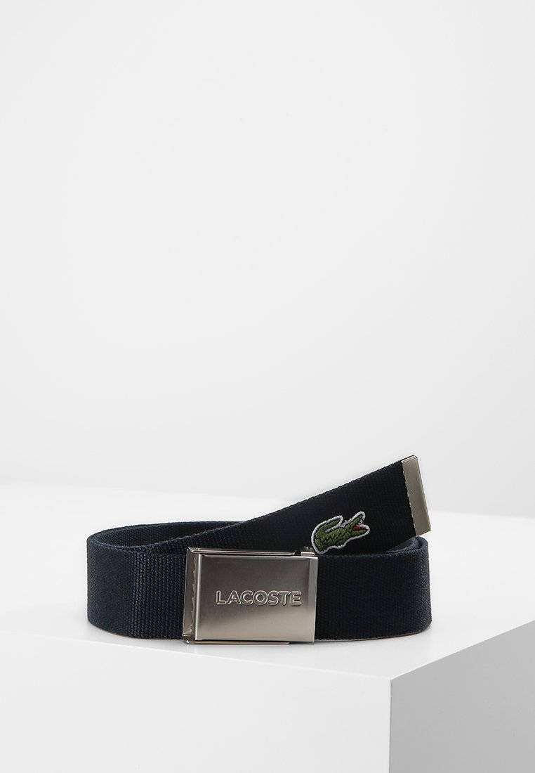 Navy fabric belt featuring a metallic rectangular buckle with engraved "LACOSTE" and a green crocodile logo embroidered on the strap.