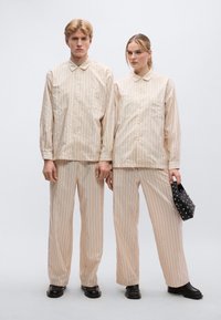 Συνολάκια μπεζ με ρίγες από ελαφρύ ύφασμα· πουκάμισα με γιακά που κουμπώνει με κουμπιά, δύο τσέπες· συνδυασμένα με φαρδιά παντελόνια· μαύρη τσάντα με διακοσμητικά καρφιά.