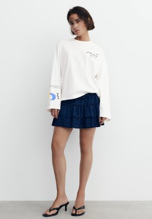 Junge Frau mit kurzen dunklen Haaren, die ein weißes Langarmshirt mit Grafik und einen marineblauen, gestuften Minirock mit schwarzen Sandaletten trägt.