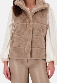 Chaleco de pelaje sintético en beige, con collar, cierre frontal de botones y paneles acolchados horizontales. Llevado sobre una camisa de manga larga blanca.