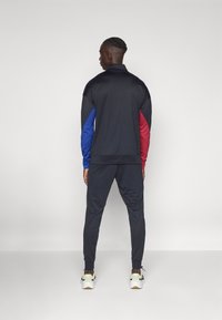 Sports tracksuit i sort, rød og blå. Har en zip-up jakke med kontrasterende ærmer og tætsiddende bukser. Lavet af letvægtsstof.