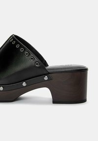 Zwarte leren klomp met grote ronde zilveren studs. Heeft een robuuste houten hak, een textuur oppervlak en een open achterkant ontwerp.