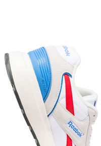Sneaker bianca con accenti blu e rossi, caratterizzata da una finitura in camoscio, tomaia in rete, suola in gomma texturizzata e etichetta "Reebok GL 1100".
