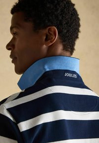 Homme aux cheveux courts et bouclés portant un T-shirt rayé bleu marine et blanc avec un col bleu clair relevé, affichant le texte "JOULES" au dos.