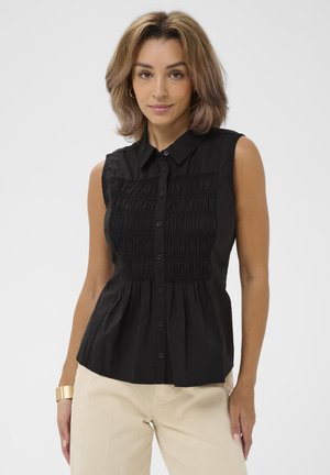 Kobieta o włosach do ramion, ubrana w bezrękawową czarną bluzkę na guziki z marszczonym przodem oraz beżowe spodnie, na jednolitym tle.