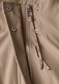 Beige interiér bundy s stříbrným zipem a logovanou táhlicí, obklopený kovovými patentkami a hladkou texturou látky.