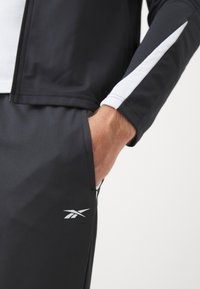 Gros plan sur une personne portant un pantalon de sport noir avec un logo blanc et une veste noire avec une bande blanche sur la manche, main dans la poche.