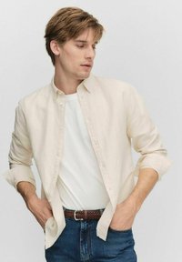 Lichtbeige overhemd met knopen over een wit T-shirt, gecombineerd met blauwe spijkerbroek. Het overhemd heeft lange mouwen die bij de manchetten zijn opgerold en een klassieke kraag.