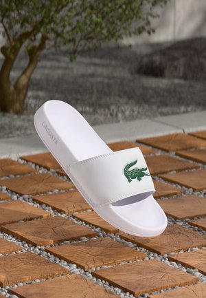 Sandale blanche à glissière avec un logo crocodile vert, fabriquée en matériau synthétique, dotée d'une large sangle et d'une semelle texturée, flottant au-dessus de carreaux à motif bois.