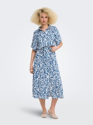 Femme aux cheveux blonds bouclés portant une robe à motifs bleus et blancs avec boutons, manches courtes et des ballerines tissées beige.