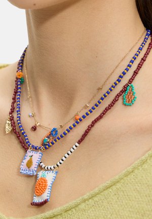 Warstwowe naszyjniki z koralików w kolorach niebieskim, bordowym i białym z haftowanymi owocowymi zawieszkami noszone przez osobę w jasnozielonym topie.