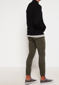 Schwarzer Sweatshirt mit Rundhalsausschnitt, graues Lagenhemd darunter, olivgrüne tapered Hose und graue Sneakers mit weißen Sohlen.