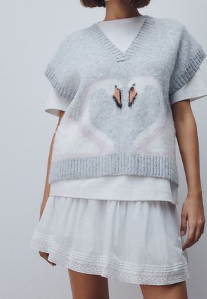 Femme portant un gilet tricoté gris clair avec deux cygnes face à face sur une chemise blanche à manches courtes et une jupe blanche.