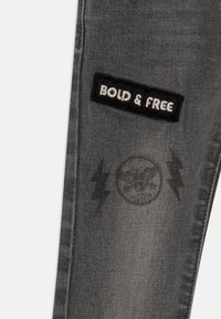 Vaqueros de mezclilla gris con un parche negro que dice "BOLD & FREE" y un gráfico redondo desgastado con rayos, que muestra una textura desgastada.