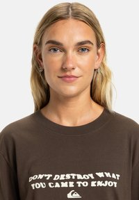 T-shirt in cotone marrone con maniche corte, che presenta una scritta bianca che recita "NON DISTRUGGERE CIÒ CHE SEI VENUTO A GODERE" e un piccolo logo.