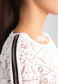Top bianco in pizzo con intricati motivi floreali, cerniera nera lungo la spalla e una texture del tessuto morbida.