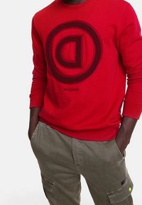 Sudadera roja con un logo circular negro que presenta una letra "D" destacada en el centro. La tela parece suave y tiene un cuello redondo.