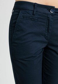 Primo piano di una persona che indossa pantaloni blu scuro con un bottone e una tasca a fessura sul lato sinistro anteriore.