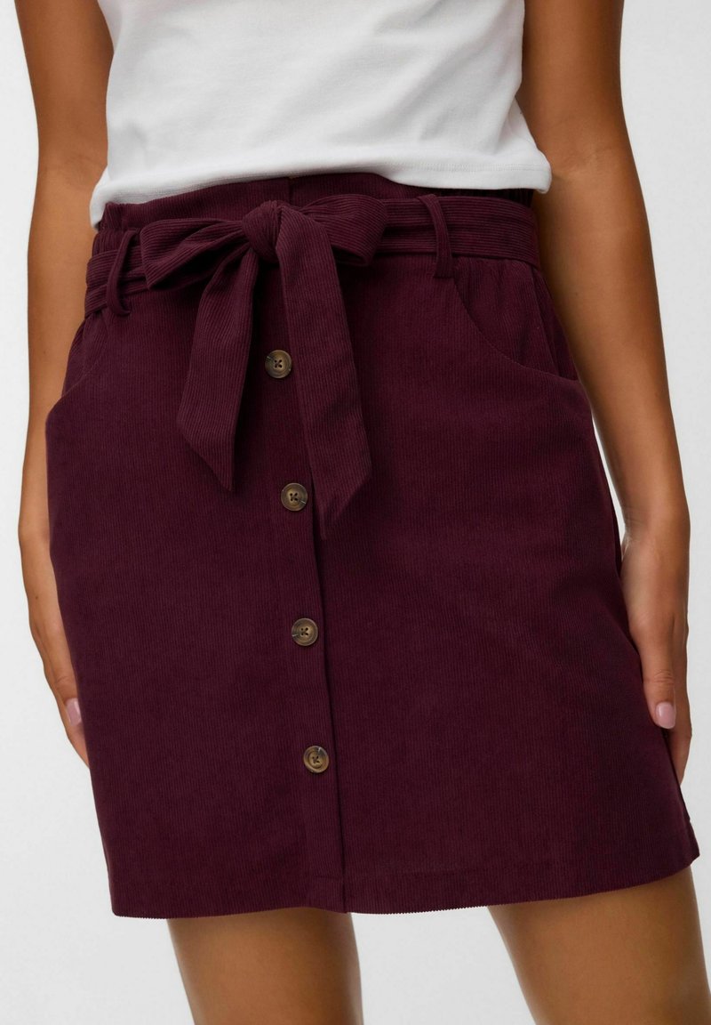 Femme portant une jupe en velours côtelé bordeaux avec boutons sur le devant, ceinture nouée à la taille et poches latérales, associée à un haut blanc.