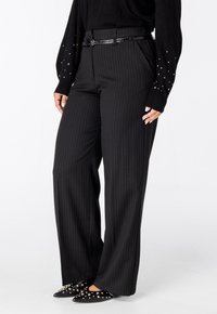 Zwarte, gestreepte wijde broek met een getailleerde pasvorm, gecombineerd met zwart puntig neuschchoenen versierd met metalen studs.