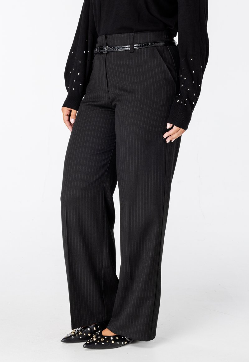 Zwarte, gestreepte wijde broek met een getailleerde pasvorm, gecombineerd met zwart puntig neuschchoenen versierd met metalen studs.