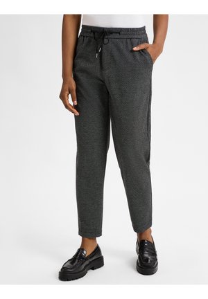 Persona vistiendo pantalones ajustados de cordón con patrón negro y gris con mocasines negros, una mano en el bolsillo, sobre un fondo blanco liso.