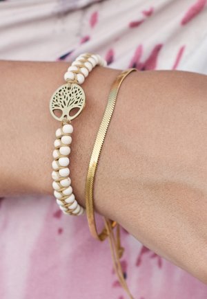 Bracciale di perline bianche con un ciondolo a forma di albero della vita verde, abbinato a un sottile bracciale dorato con una finitura testurizzata.