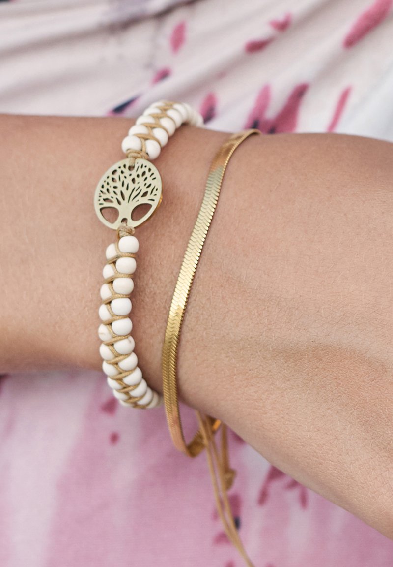 Weißes Perlenarmband mit einem grünen Baum-des-Lebens-Anhänger, kombiniert mit einem dünnen goldenen Armreif mit strukturierter Oberfläche.