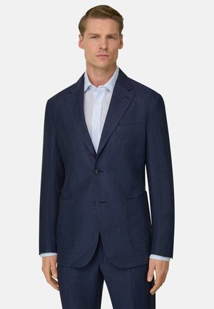 Giovane uomo che indossa un abito blu scuro con una camicia elegante a righe azzurre, in piedi davanti a uno sfondo chiaro e semplice.