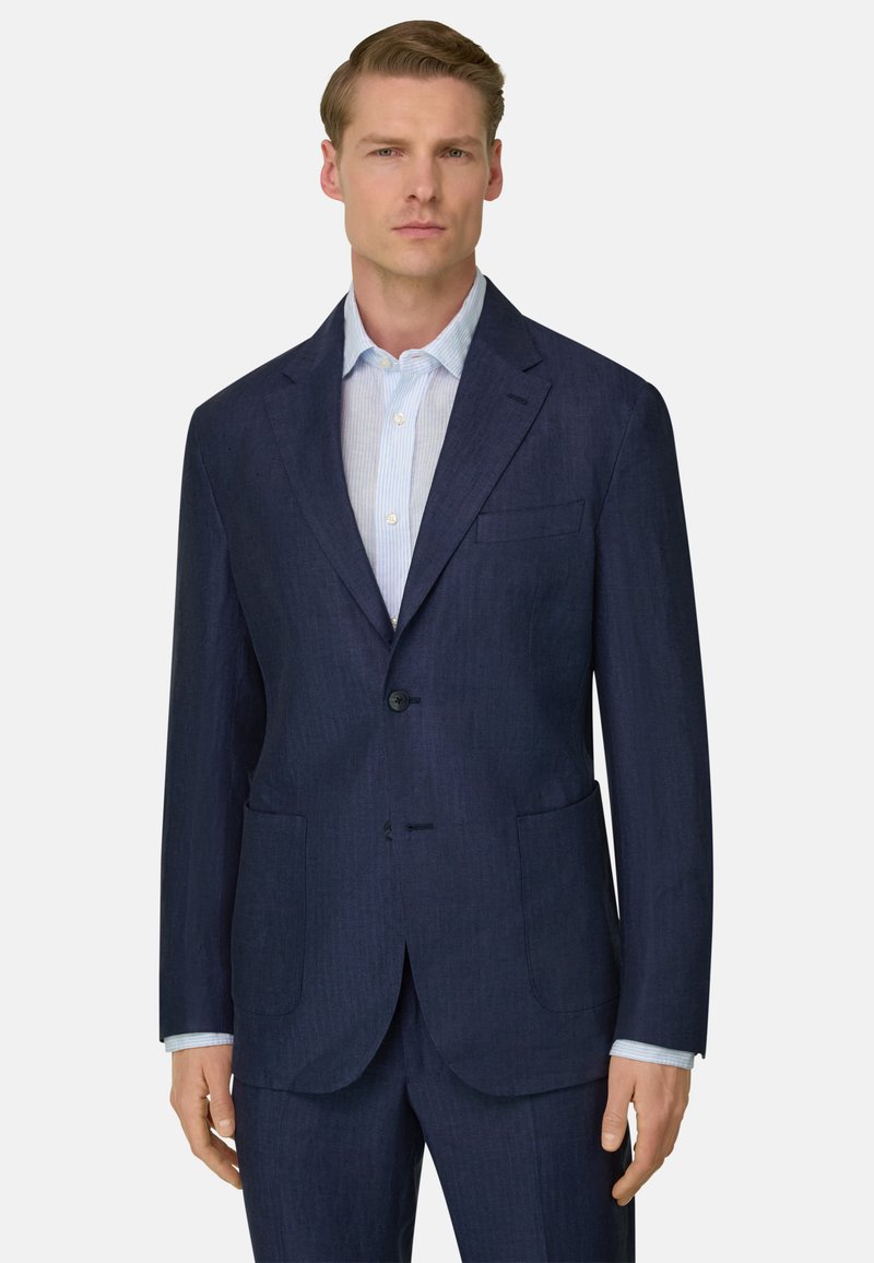 Jeune homme portant un costume bleu foncé avec une chemise à rayures bleues claires, debout devant un fond clair uni.