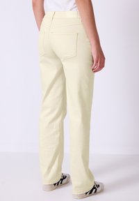 Pantalones de mezclilla amarillo claro con diseño de pierna recta, bolsillos traseros destacados y costura en contraste. Combinados con zapatillas negras y blancas.