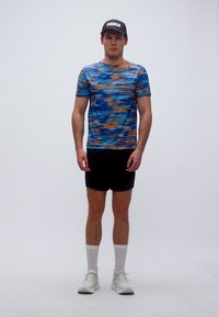 Άνδρας που στέκεται φορώντας μαύρο καπέλο HOKA, μπλε και πορτοκαλί πουκαμίσα με κοντά μανίκια και σχέδιο, μαύρο σορτς, λευκές κάλτσες και λευκά αθλητικά παπούτσια σε απλό φόντο.