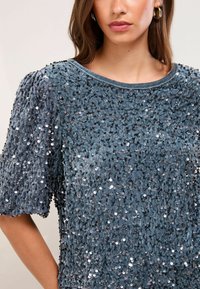 Tīrā zila samta blūze ar īsām pūkām, rotāta ar sudrabainiem spīdekļiem. Gluda tekstūra ar apaļu izgriezumu un mirdzošiem elementiem visā virsmā.