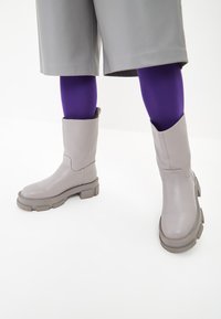 Botas de tobillo grises con un acabado de cuero suave, suela gruesa y texturizada, y un eje ancho. Llevadas con medias moradas y un atuendo gris.